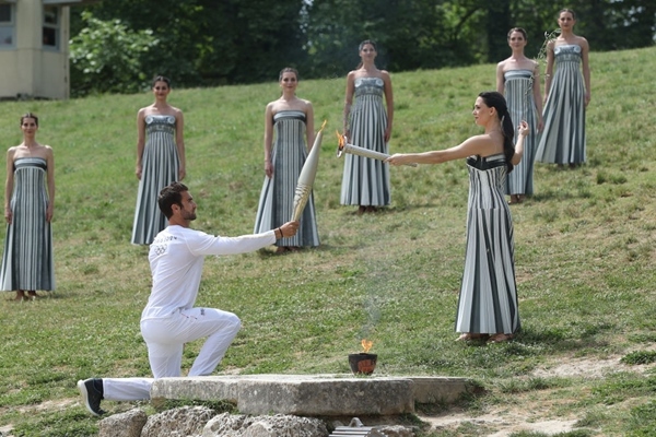 扮演最高女祭司的演員瑪麗·米娜（前右）點(diǎn)燃第一棒火炬手——希臘運(yùn)動(dòng)員、東京奧運(yùn)會(huì)男子賽艇項(xiàng)目金牌得主斯特凡諾斯·恩圖斯科斯手中的火炬。新華社記者 趙丁喆 攝